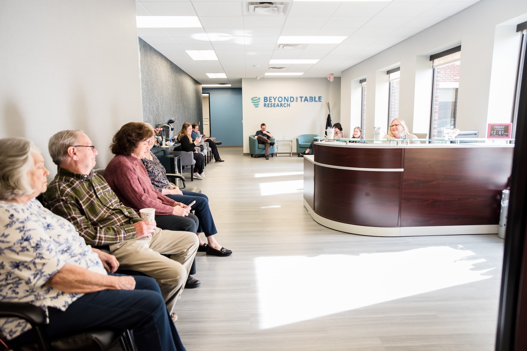 A bright, modern office reception area with a curved wooden front desk and glass countertop. Black chairs line one wall for seating, and additional gray chairs and a coffee station are visible toward the back. The space features light gray wood flooring, neutral walls with a blue accent section, and a sign reading “Beyond the Table Research” on the far wall.