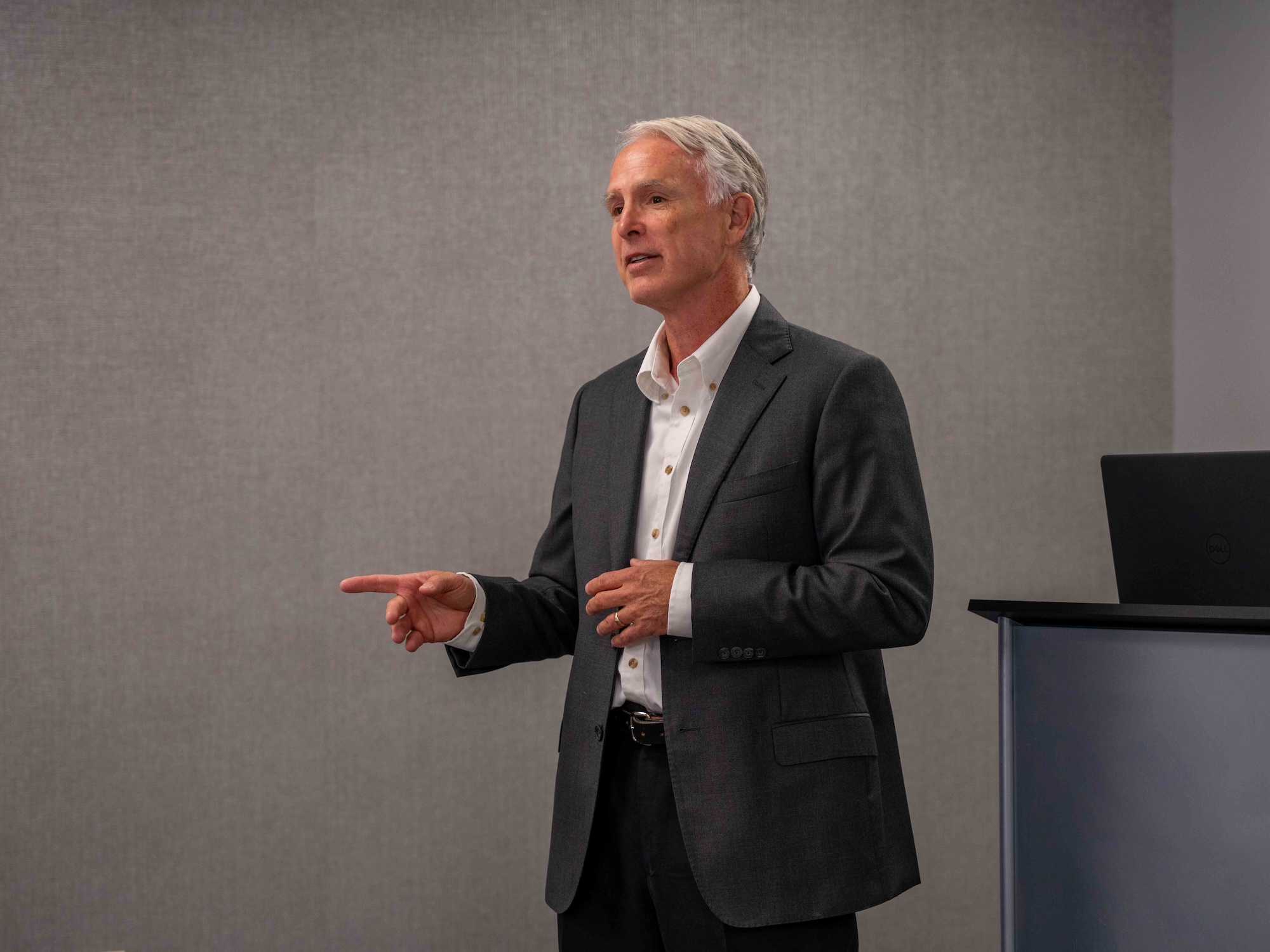 An older man in a dark suit giving a presentation, gesturing with his hand while speaking in front of a gray wall and a podium with a laptop.