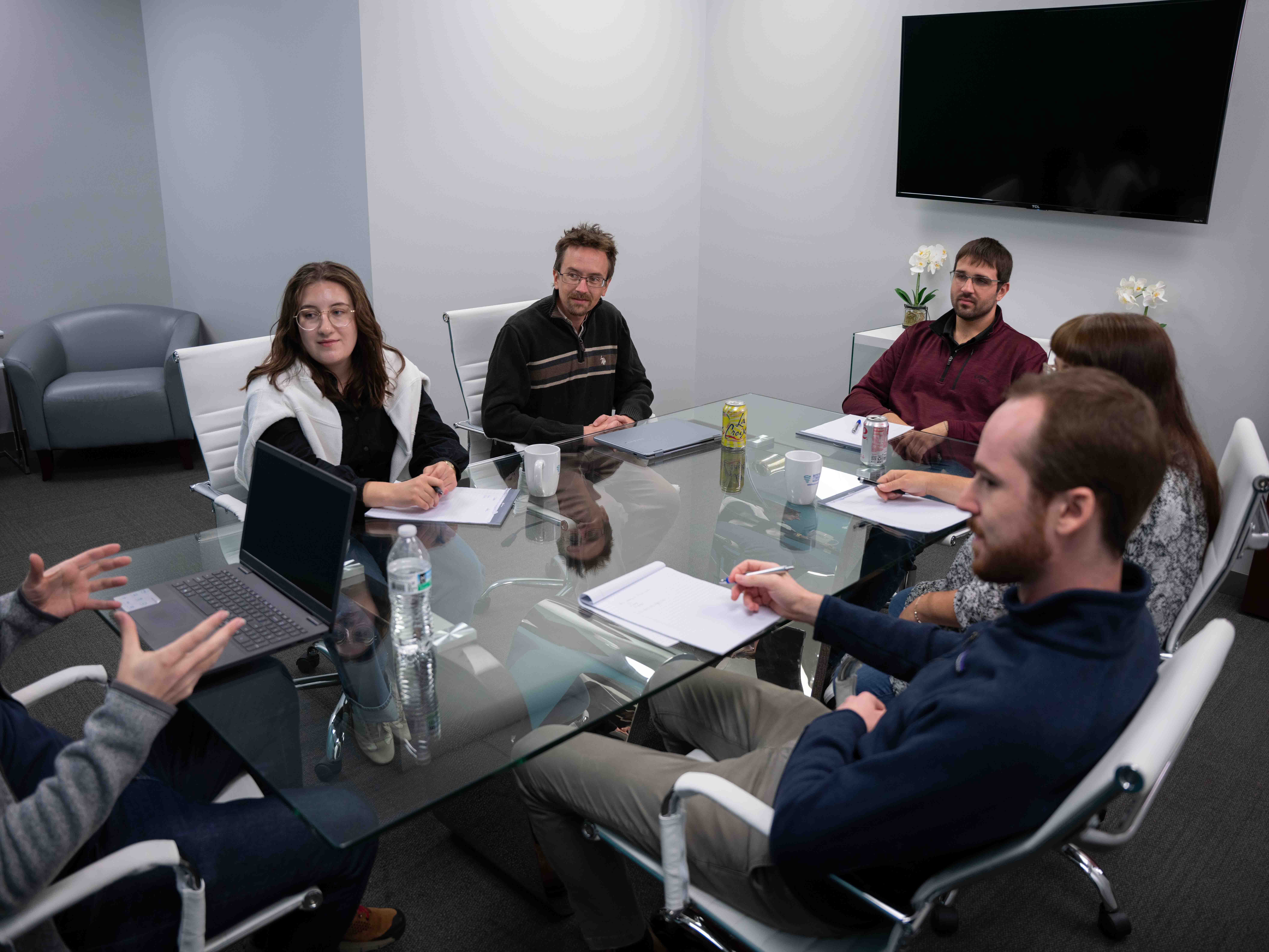 Six people sit around a glass conference table during a meeting, with laptops, notebooks, and drinks in front of them in a modern office space.